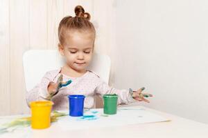 hermosa pequeño niña sorteos con dedo pinturas en un blanco sábana de papel. creativo niño desarrollo en jardín de infancia o gratis hora a hogar foto