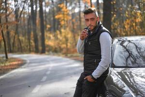 A handsome young Caucasian man wearing style black vest standing near black car on nature. photo