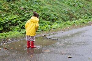 Playful girl wearing yellow raincoat while jumping in puddle during rainfall photo