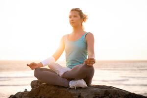 mujer práctica yoga loto actitud a meditación con verano vacaciones rocoso playa felicidad y relajación. calma hembra ejercicio con yoga meditar Oceano playa con puesta de sol dorado tiempo. foto