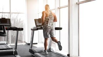 Muscular athletic bodybuilder fitness model running treadmill gym near big window photo