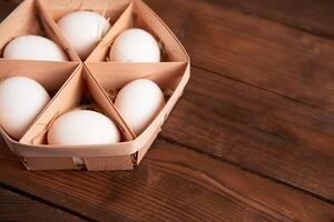 White chicken eggs lie in round wooden basket which stands on a dark wooden table. photo