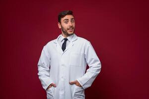 Young handsome modern doctor in a white medical gown stands in the studio on a red background. Student trainee of a medical university. photo