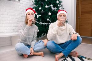 joven contento Pareja en Navidad sombreros cerca un Navidad árbol besando, participación lentes de vino. nuevo año celebracion foto