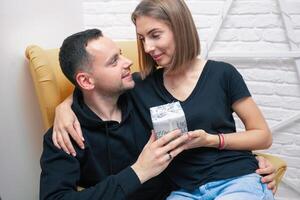 Portrait of couple young in a cozy room. A man sits on a yellow chair. A woman sits on the lap of a man and holds a gift photo