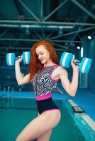 Beautiful young red-haired girl in a fashionable swimsuit in the style of the 80s stands with the dumbbells in her hands in the indoor swimming pool photo
