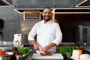 Handsome young African chef standing in professional kitchen in restaurant preparing a meal of meat and cheese vegetables. photo