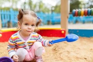 a little girl with two tails is dressed in a striped colorful jacket is playing in the sandbox on the playground photo