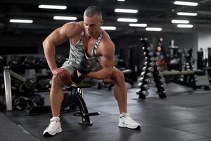 Muscular athletic bodybuilder fitness model sitting bench training biceps lift dumbbells in indoor gym photo