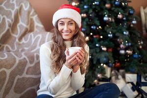 Christmas. Woaman dressed white sweater Santa hat and jeans sitting on the floor near christmas tree with present box photo