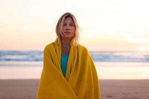 Beautiful young woman wrapped in yellow blanket standing on sandy beach at sunset looking camera. Beautiful caucasian female person wrapped cozy plaid standing alone on ocean beach photo