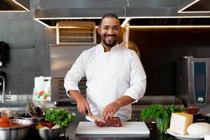 Handsome young African chef standing in professional kitchen in restaurant preparing a meal of meat and cheese vegetables. photo