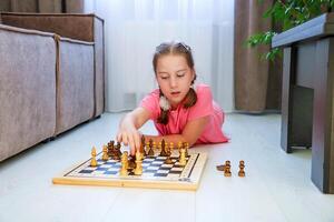 pequeño niña a hogar acostado en el piso haciendo se mueve con piezas en el tablero de ajedrez foto