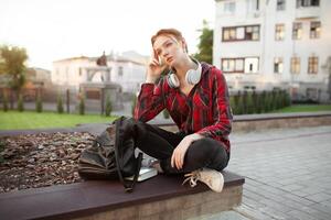 A red-haired girl with freckles is dressed in a red checked shirt is sitting in a lotus pose in the park listening to music on headphones Modern student photo