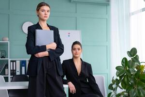 Two young women leaders dressed black suit in office one woman standing with document folder other woman sitting on the table looking at camera. Business meeting photo