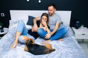 Young couple sitting on a bed in the bedroom, hugging. Near them is their dog photo