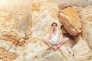 un hermosa joven niña con corto pelo es vestido en pantalones cortos y un blanco jersey es practicando yoga en el antecedentes de rocas actitud de el loto. el concepto de calma y concentración foto