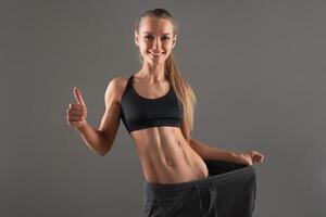 Full-length portrait of attractive slim young smiling woman in big pants showing successful weight loss with her thumb up, isolated on gray background photo