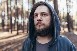 un joven hombre con un barba camina en un pino bosque. retrato de un brutal barbado hombre en un capucha foto