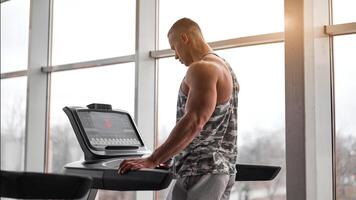 Muscular athletic bodybuilder fitness model running treadmill gym near big window photo