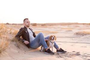 un joven caucásico hombre vestido negro cuero chaqueta y azul pantalones se sienta en arenoso playa siguiente a su amigo el perro beagle criar. foto