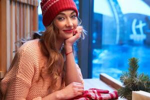 Beautiful caucasian blonde woman in red knitted hat sitting behind cafeteria window drinking a warm latte on a wonderful Christmas day. photo