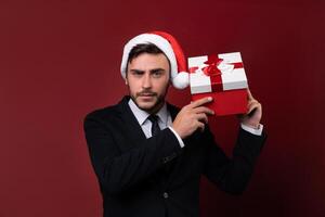 joven hermoso caucásico chico en negocio traje y Papa Noel sombreros soportes en rojo antecedentes en estudio y carita sostiene cerca el oído rojo regalo caja foto