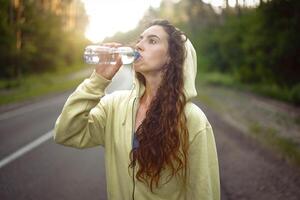 Beautiful young Caucasian girl  standing on asphalt forest road Drinks water from plastic bottle after jogging photo