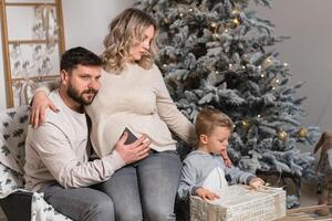 Navidad familia felicidad retrato de papá, embarazada mamá y pequeño hijo sentado Sillón a hogar cerca Navidad árbol abrazo sonrisa foto