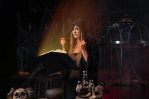 Halloween concept. Witch dressed black hood with dreadlocks standing dark dungeon room use magic book for conjuring magic spell photo