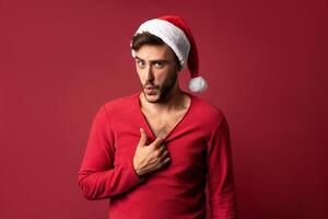 Young handsome caucasian guy in red sweater and Santa hats stands on red background in studio  Shows how hot he is photo