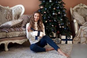 Christmas. Woman dressed white sweater and jeans sitting on the floor near christmas tree with present box photo