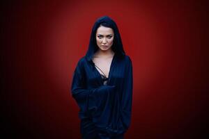 Young beautiful woman with a black hair and in the dark blue cloak with hood at the red background photo