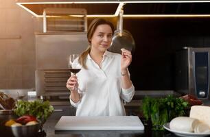 Beautiful young caucasian girl standing in kitchen in a white uniform smiling and tasting red wine photo