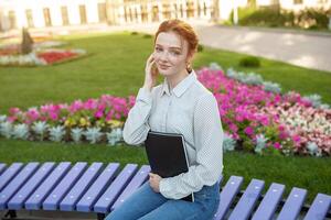 Young beautiful red-haired girl with freckles sitting on a bench near the university holds a notebook in her hands with homework. Portrait of a student. Back to school concept. photo