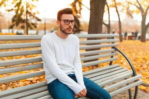 A lonely handsome man sits sad in the park on a bench. Autumn season, yellow leaves on background. Loneliness concept. The student did not pass the exams or broke up with the girl photo