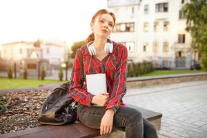 young beautiful student wearing a checkered shirt headphones and a backpack resting on the street between classes photo