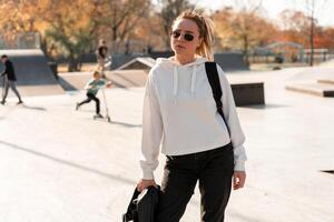 Outdoor portrait of young beautiful woman with a ponytail and sunglasses, with a backpack on his shoulders, dressed in a white sweater, near the sportsground photo