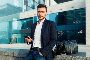 Handsome young fashionable man in a stylish business suit and sunglasses is typing a message in his phone walking in the autumn park on the background of bright yellow leaves. photo