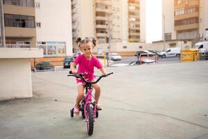 Cute little smiling girl riding bike bicycle in city on parking sunny summer day. Active family leisure with kids. photo