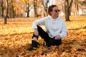 Woman in a white sweater with a hood sits on ground in the park and holding a leaf in her hands photo