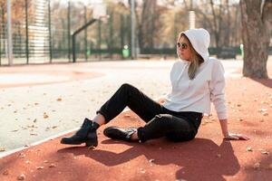 al aire libre retrato de joven hermosa mujer con largo en Gafas de sol y un blanco encapuchado suéter sentado en el campo deportivo pista foto