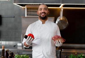 contento sonriente cocinero prepara carne plato con varios vegetales en el cocina. en uno mano el hombre sostiene verduras, en el otro un pedazo de Fresco carne foto