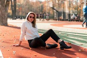 al aire libre retrato de joven hermosa mujer con largo en Gafas de sol y un blanco encapuchado suéter sentado en el campo deportivo pista foto