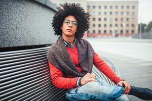 A handsome young Egyptian curly guy sitting on a bench dressed in a bright orange sweater and jeans. The student walks in the break between the lessons. Cute hipster guy photo