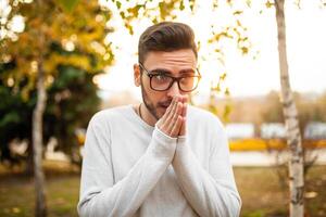 Young handsome hipster man in glasses and a white sweater is walking in a cold autumn park. Heats his hands with his breath. Concept of seasonal autumn cold photo