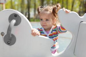 un pequeño niña con dos cruz es vestido en un a rayas vistoso chaqueta es jugando en el patio de recreo foto