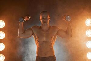 Young pumping dumbbells, practicing biceps training, smoky illuminated background photo