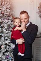 alegre Navidad y contento nuevo año. papá en un negocio traje sostiene su hija vestido en un elegante rojo vestir en el antecedentes de el Navidad árbol foto