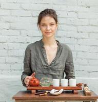 Oriental Master of tea ceremony with white brick wall on the background. Traditional tea party on the nature with woman dressed in the gray shirt photo
